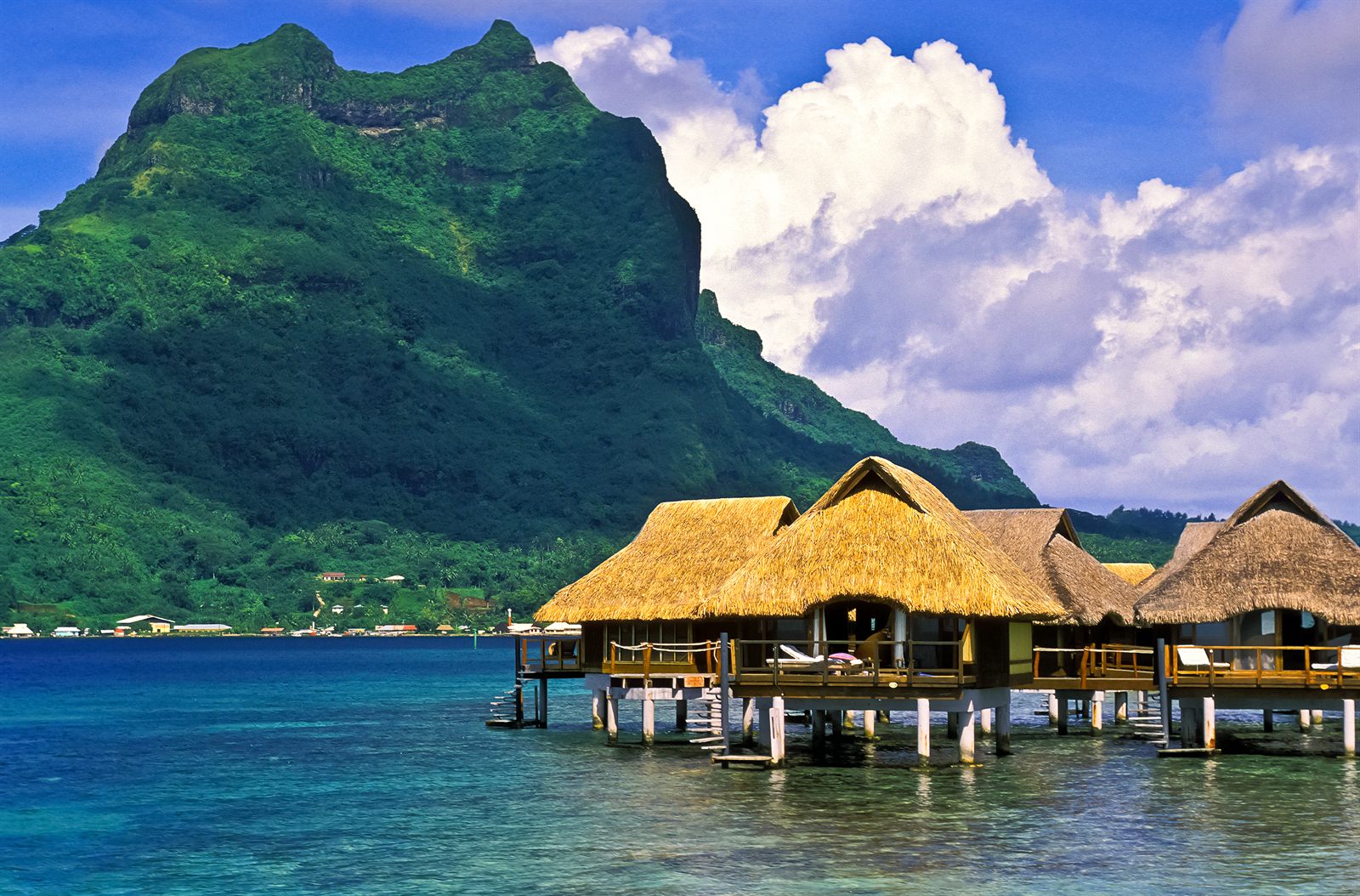 Thatched overwater bungalows below a green volcanic peak in French Polynesia